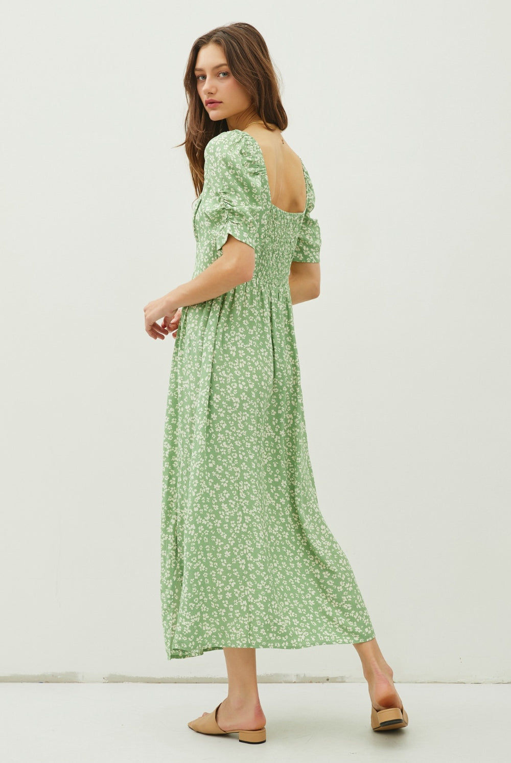 A poised woman exhibits a serene and romantic look in a sage green maxi dress with a delicate floral print and a tasteful back slit, perfect for spring and summer occasions.