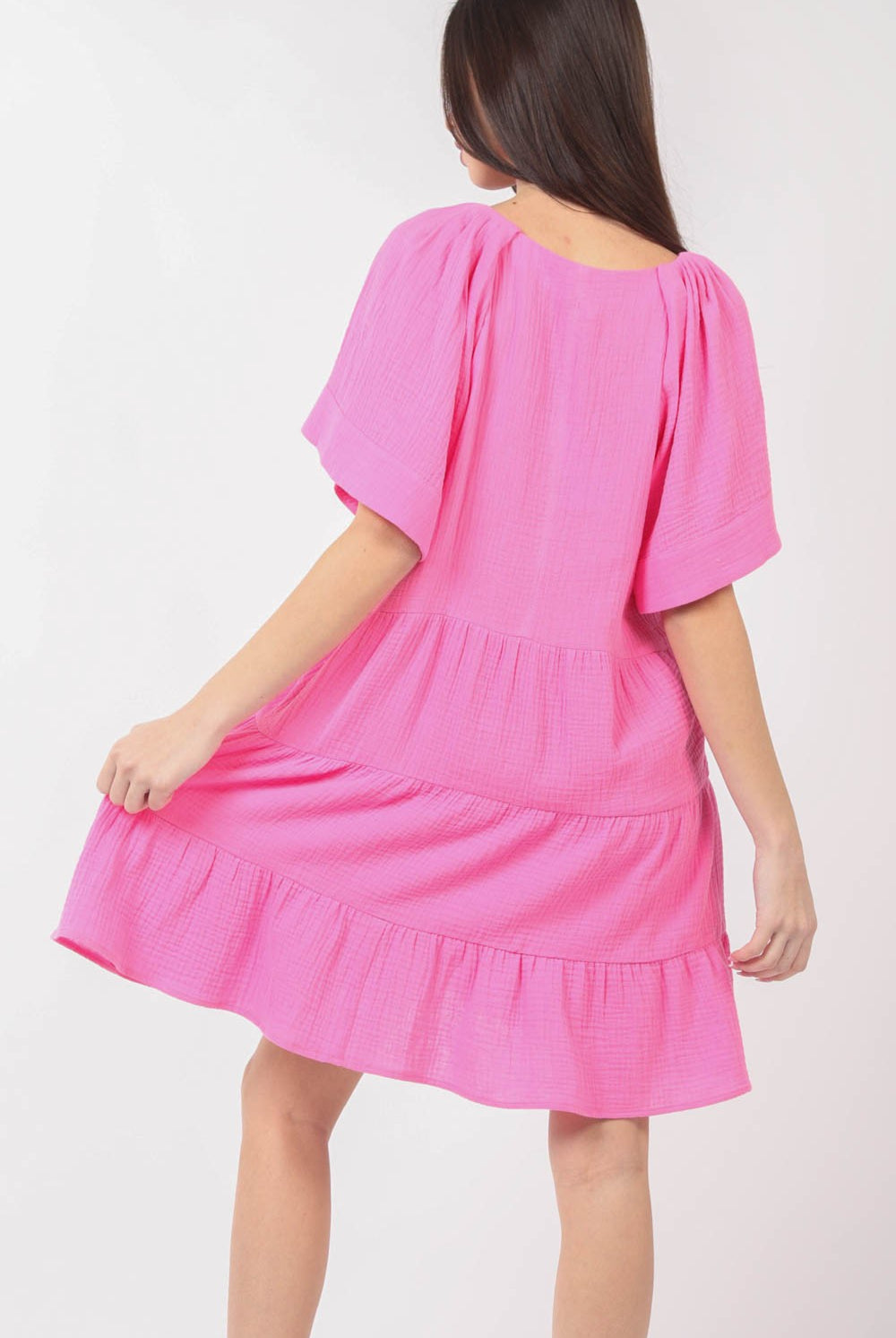 A model wears a vibrant pink, ruffled tiered short dress with a V-neckline, flutter sleeves, and a flounce hem, paired with white sandals with ruffle details.