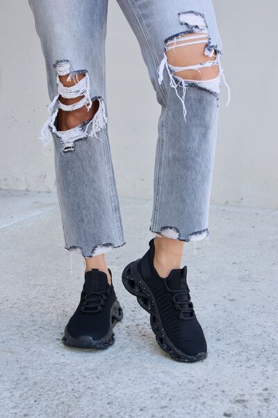 Black athletic sneakers with unique bubble soles and speckled details, paired with frayed gray cropped jeans, against a concrete surface.