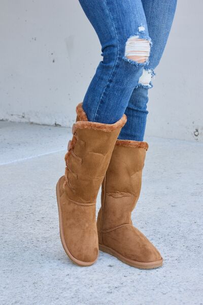A pair of women's mid-calf fur-lined boots in a tan suede material, featuring side button details and a patterned sole, paired with blue denim jeans.