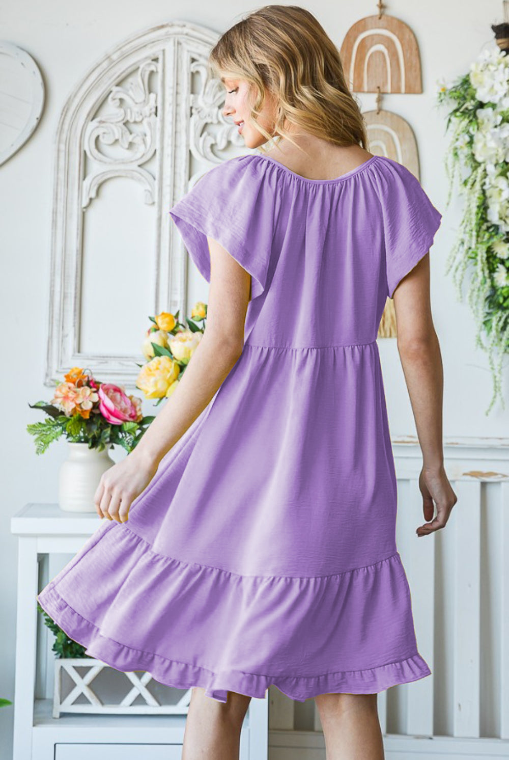 A woman in a summery setting wears a light lavender short sleeve dress with a ruffle hem, complemented by a wide-brimmed straw hat, embodying a fresh, spring-ready vibe.