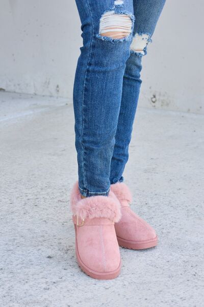 Pink fur-lined ankle boots with a gold buckle detail, paired with ripped blue skinny jeans, presented against a concrete background.