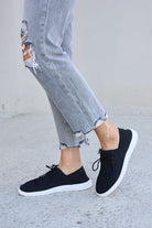 Black mesh sneakers with white soles and black laces, worn with frayed gray ankle-length jeans, against a concrete background.