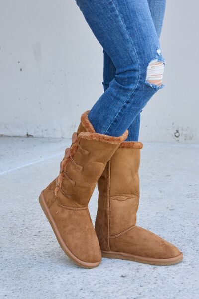 A pair of women's mid-calf fur-lined boots in a tan suede material, featuring side button details and a patterned sole, paired with blue denim jeans.