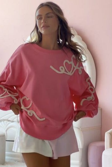 A model in a relaxed pose wearing a white round neck sweatshirt with pink 'XOXO' graphic text, paired with pink denim shorts, against a pink and white patterned background.