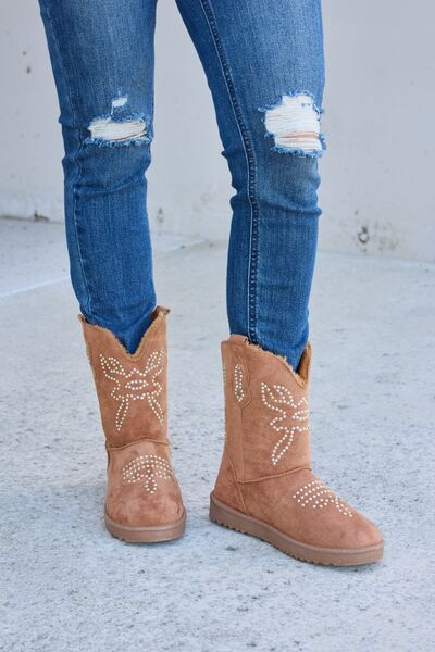 A pair of tan suede ankle boots with decorative white floral beadwork, paired with blue skinny jeans, against a simple concrete background.