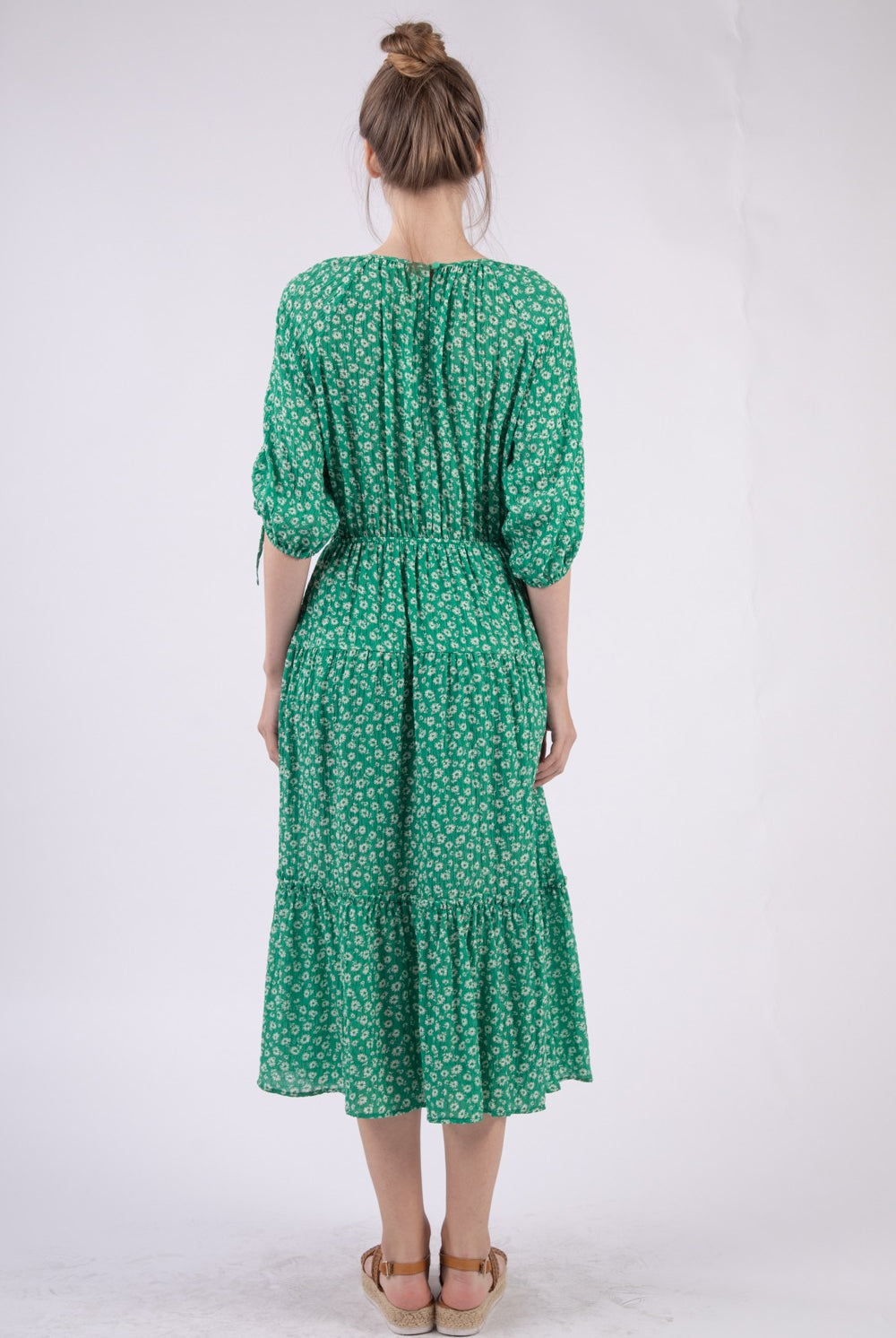 A poised woman in a fresh green midi dress adorned with a small white floral print. The dress features a cinched elastic waist, tie-up details at the three-quarter length sleeves, and a tiered skirt that adds movement. She pairs the look with natural brown strappy wedge sandals, perfect for a breezy day out.