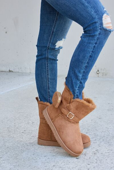A pair of tan ankle-high boots with faux fur trim and a decorative side buckle, set against a concrete background, complemented by distressed blue skinny jeans.