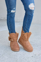 A pair of tan ankle-high boots with faux fur trim and a decorative side buckle, set against a concrete background, complemented by distressed blue skinny jeans.