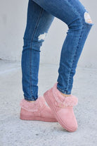 Pink fur-lined ankle boots with a gold buckle detail, paired with ripped blue skinny jeans, presented against a concrete background.