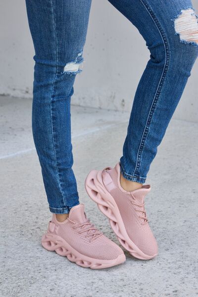 Blush pink knit athletic shoes with matching sculpted soles, paired with cuffed blue distressed jeans, on a concrete background.