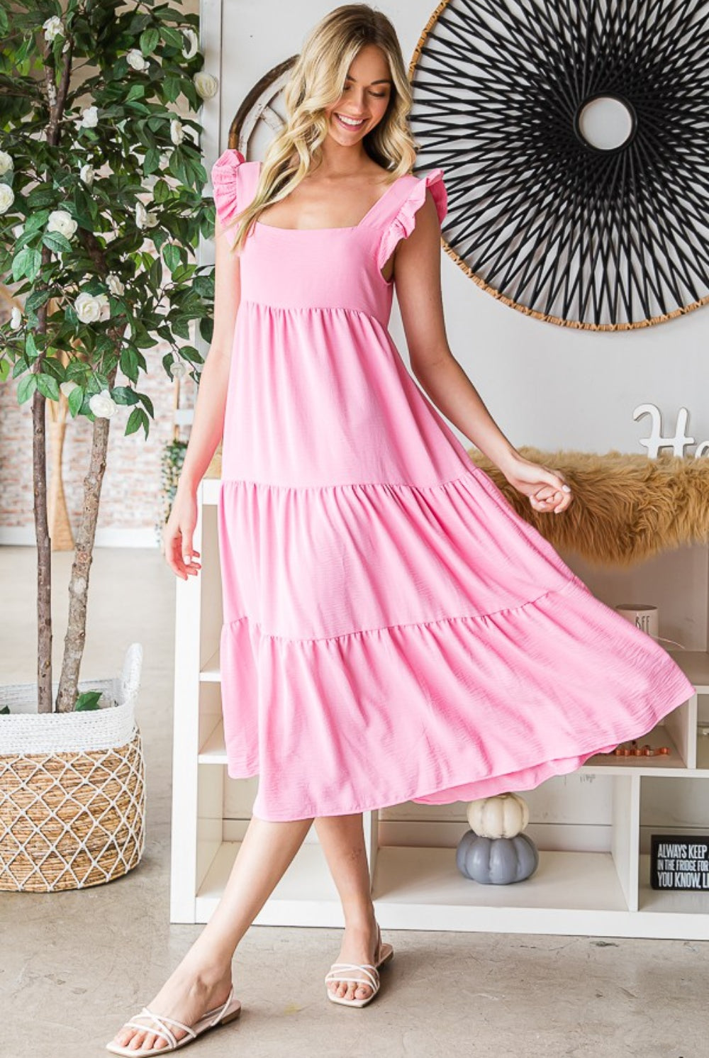 A smiling woman looks radiant in a blush pink ruffle tiered midi dress, which exudes summer charm with its flouncy sleeves and flowing skirt, complemented by white slide sandals.