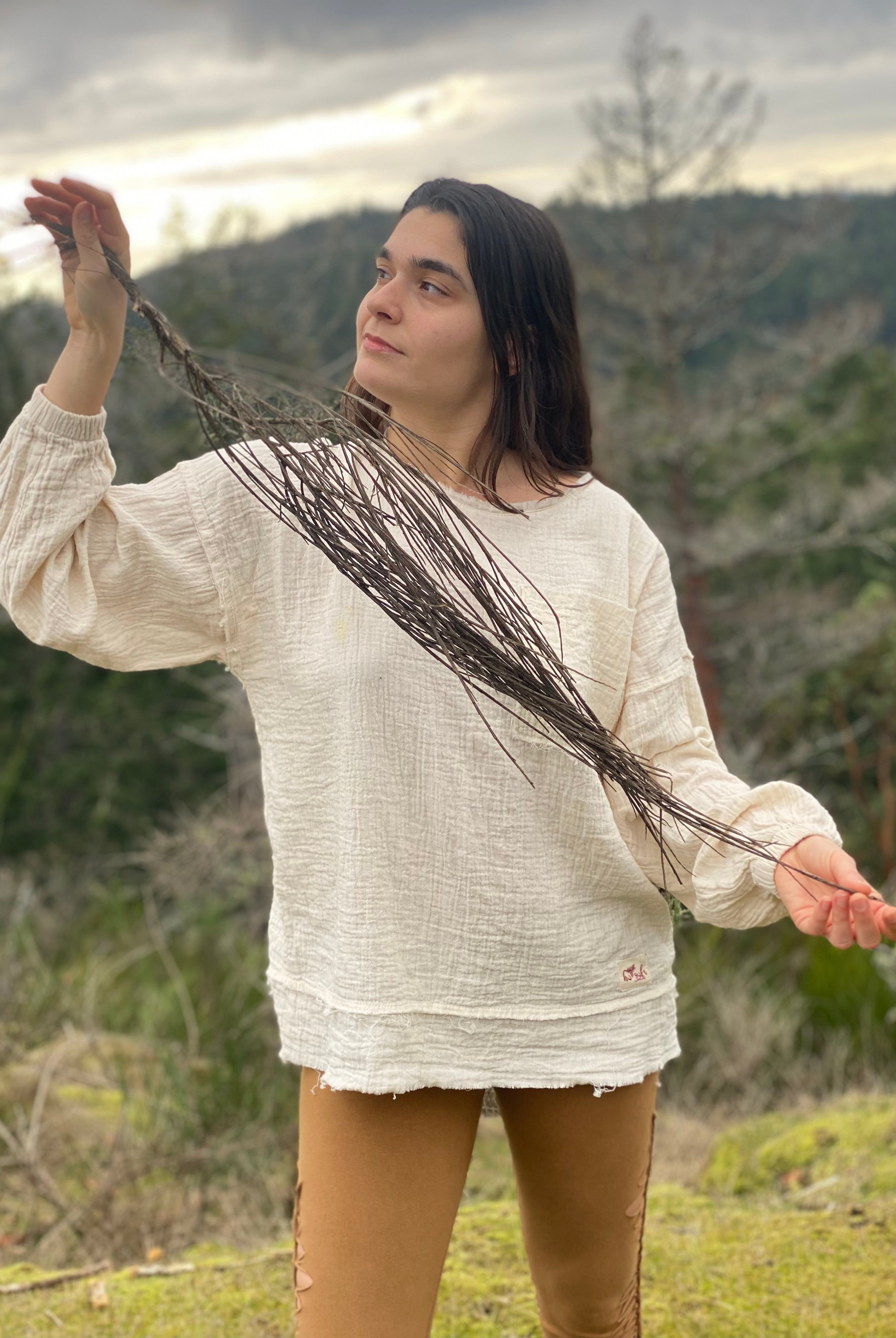 This image shows a woman in a natural outdoor setting, enjoying a moment of tranquility. She is dressed in an off-white, textured, long-sleeve blouse with a relaxed fit, paired with form-fitting, olive-green leggings and appears to be holding a thin branch or twig, eyes closed, seemingly appreciating the scent or texture of nature's offering.
