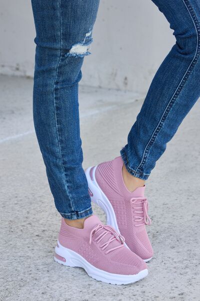 Dusty pink knit athletic shoes with white soles, paired with cuffed blue jeans featuring distressed details, on a concrete background.