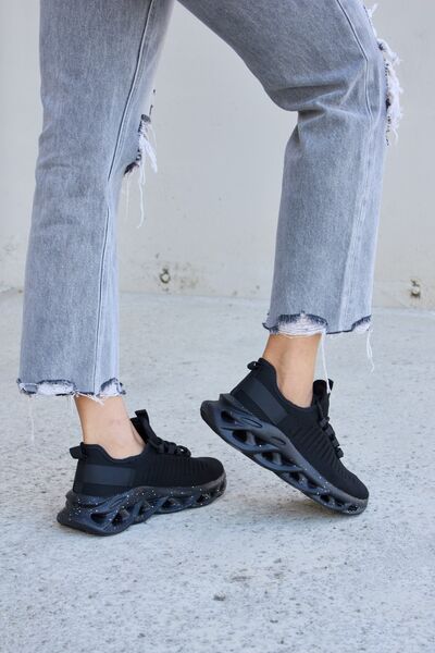 Black athletic sneakers with unique bubble soles and speckled details, paired with frayed gray cropped jeans, against a concrete surface.