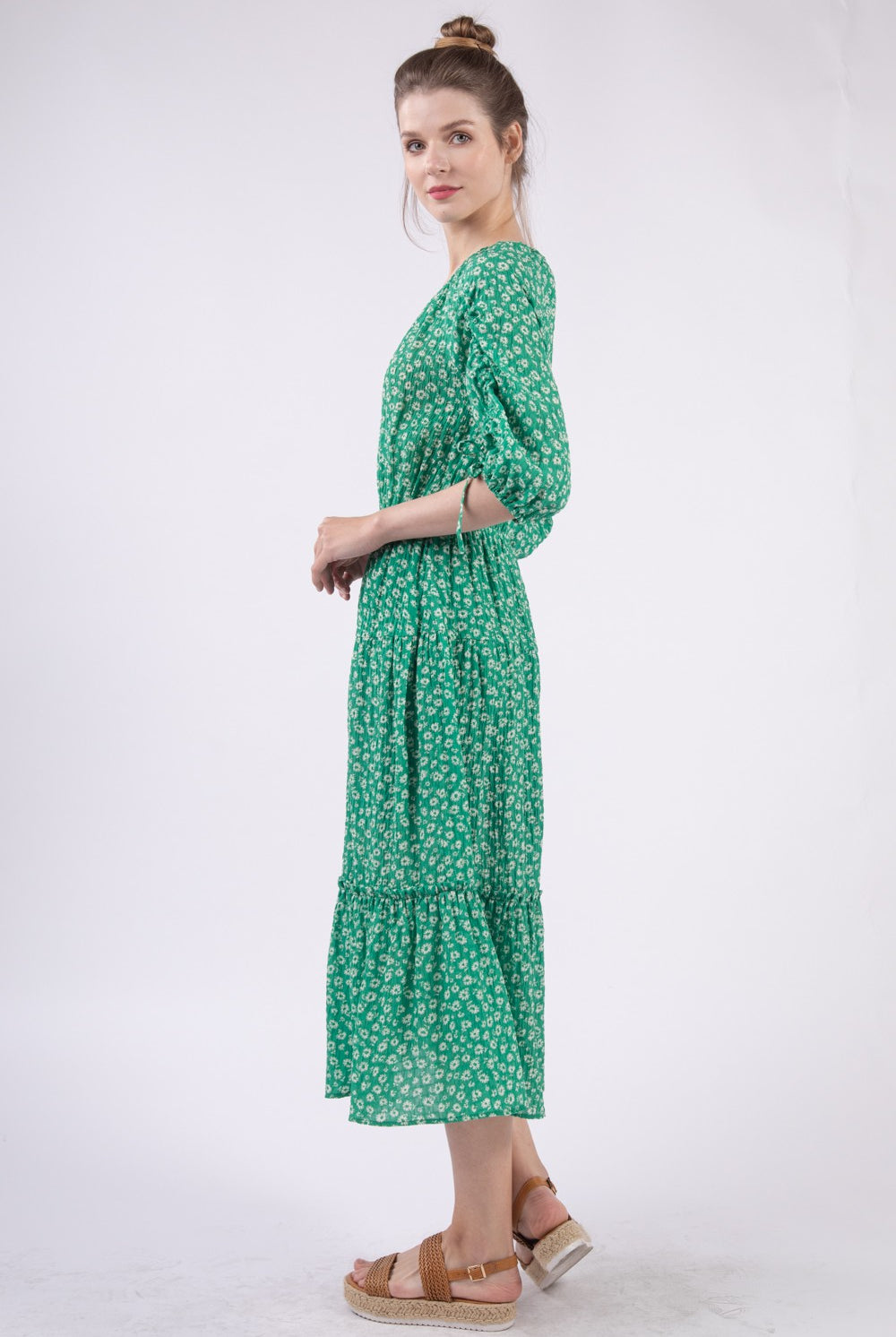 A poised woman in a fresh green midi dress adorned with a small white floral print. The dress features a cinched elastic waist, tie-up details at the three-quarter length sleeves, and a tiered skirt that adds movement. She pairs the look with natural brown strappy wedge sandals, perfect for a breezy day out.