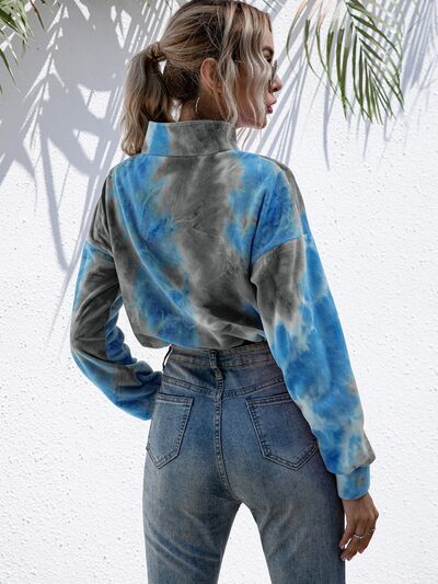 Woman in a tie-dye quarter-zip sweatshirt with drawstring hem, paired with distressed jeans and accessorized with round sunglasses, in a tropical setting with palm shadows.