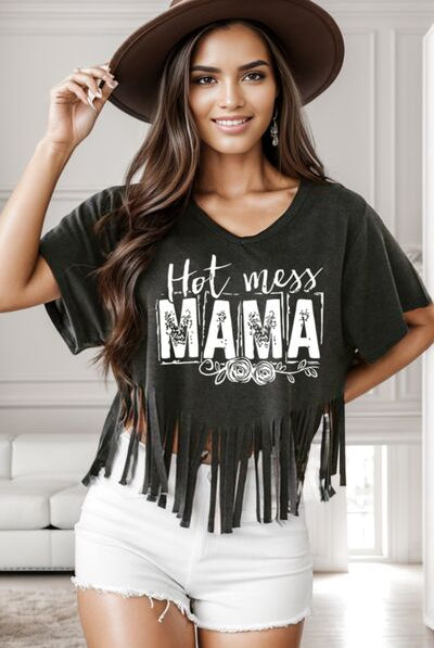 Woman posing in a casual graphic t-shirt with the phrase "Hot Mess Mama" and fringe detailing, paired with white shorts and accessorized with a wide-brimmed hat.