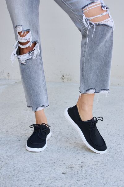 Black mesh sneakers with white soles and black laces, worn with frayed gray ankle-length jeans, against a concrete background.