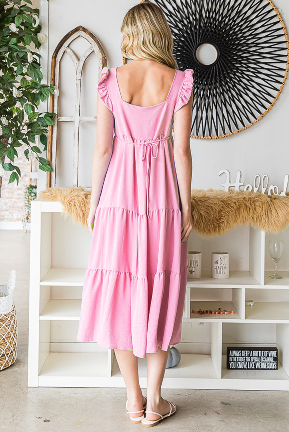 A smiling woman looks radiant in a blush pink ruffle tiered midi dress, which exudes summer charm with its flouncy sleeves and flowing skirt, complemented by white slide sandals.