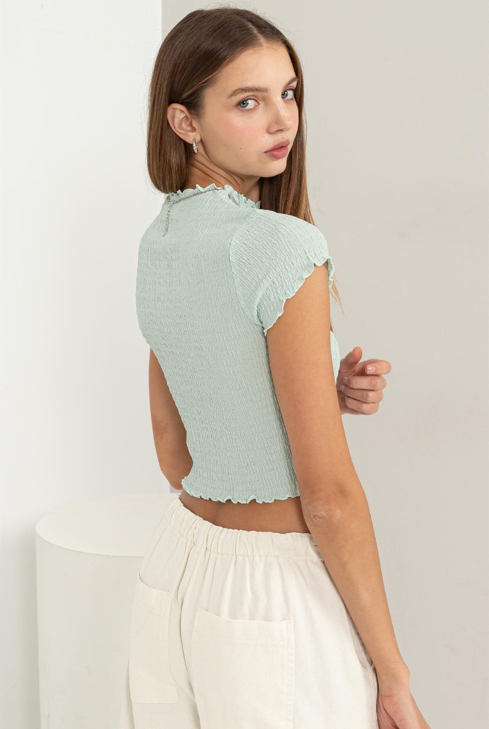 A woman models a light blue HYFVE Lettuce Hem Crop Top, paired with casual white drawstring pants. The top features short sleeves and a textured, ruffled hemline that adds a playful touch to the fitted silhouette. The image is part of an Instagram post for GemThreads Boutique to showcase their latest summer fashion item, inviting viewers to visit their website and shop the collection.