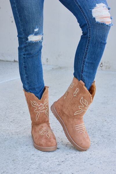 A pair of tan suede ankle boots with decorative white floral beadwork, paired with blue skinny jeans, against a simple concrete background.
