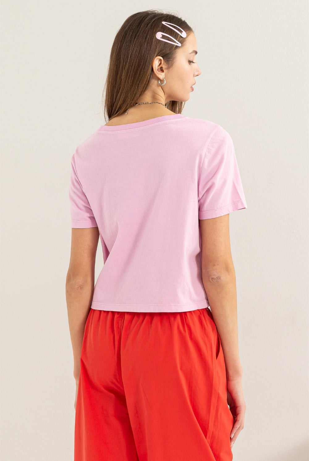 A model showcases a pastel pink HYFVE Round Neck Cropped T-Shirt, paired with vibrant red trousers. The tee has a relaxed fit, short sleeves, and a classic round neckline, exemplifying casual comfort with a touch of modern style. This image is presented to feature the product available for purchase at GemThreads Boutique, highlighting the garment's color and design for potential customers.
