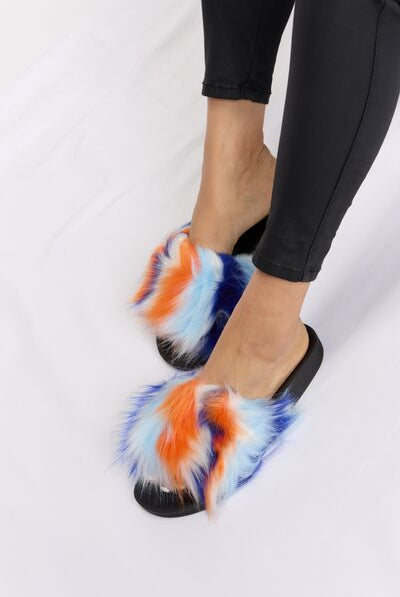 Colorful faux fur sandals with orange, blue, and white tones on a black base, paired with black ankle-length pants, against a white satin backdrop.