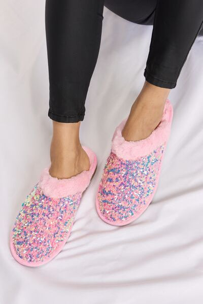 Cozy pink slippers with a multicolored sequin upper and soft faux fur lining, paired with black leggings, on a white satin background.