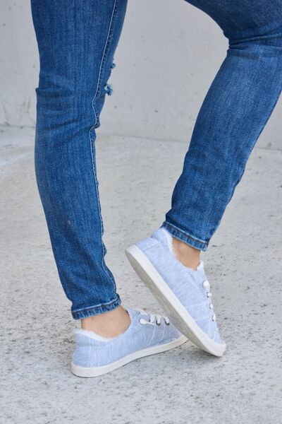Light blue denim canvas sneakers with white laces and soles, worn with cuffed blue jeans, on a concrete surface.