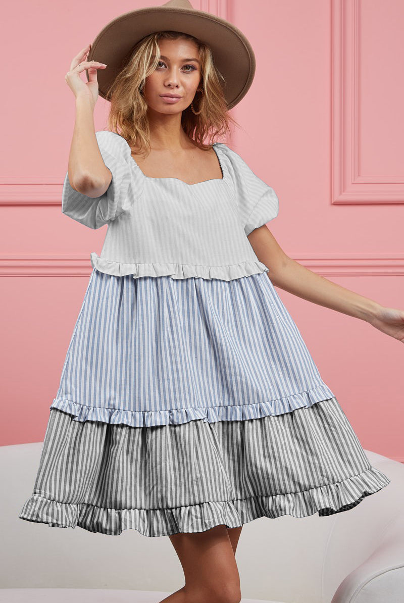 A joyful woman poses in a playful ruffled tiered mini dress with light blue and white vertical stripes, featuring flutter sleeves and a square neckline. She complements the dress with a wide-brimmed tan hat, creating a fresh and fashionable summer look against a pink background.