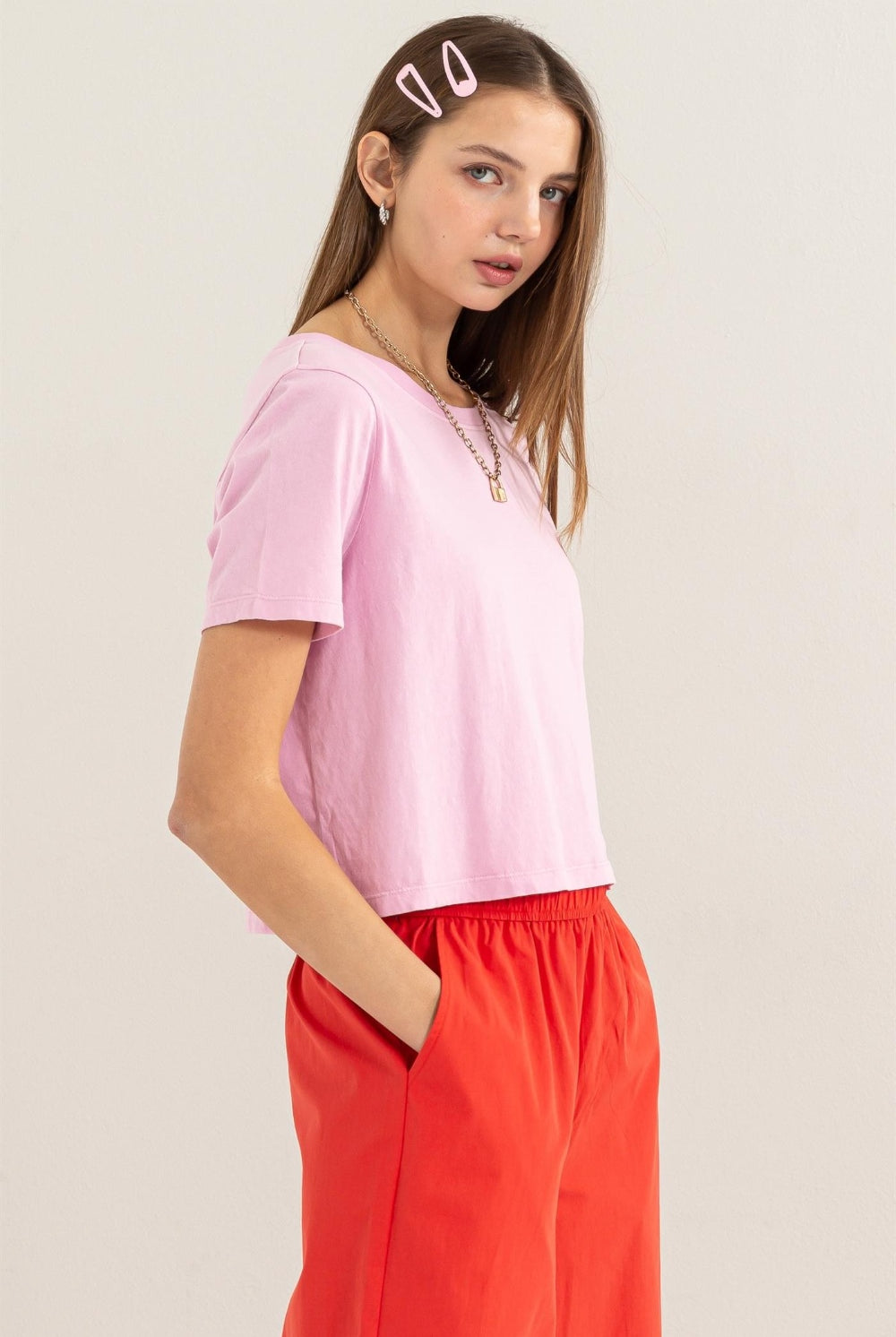 A model showcases a pastel pink HYFVE Round Neck Cropped T-Shirt, paired with vibrant red trousers. The tee has a relaxed fit, short sleeves, and a classic round neckline, exemplifying casual comfort with a touch of modern style. This image is presented to feature the product available for purchase at GemThreads Boutique, highlighting the garment's color and design for potential customers.