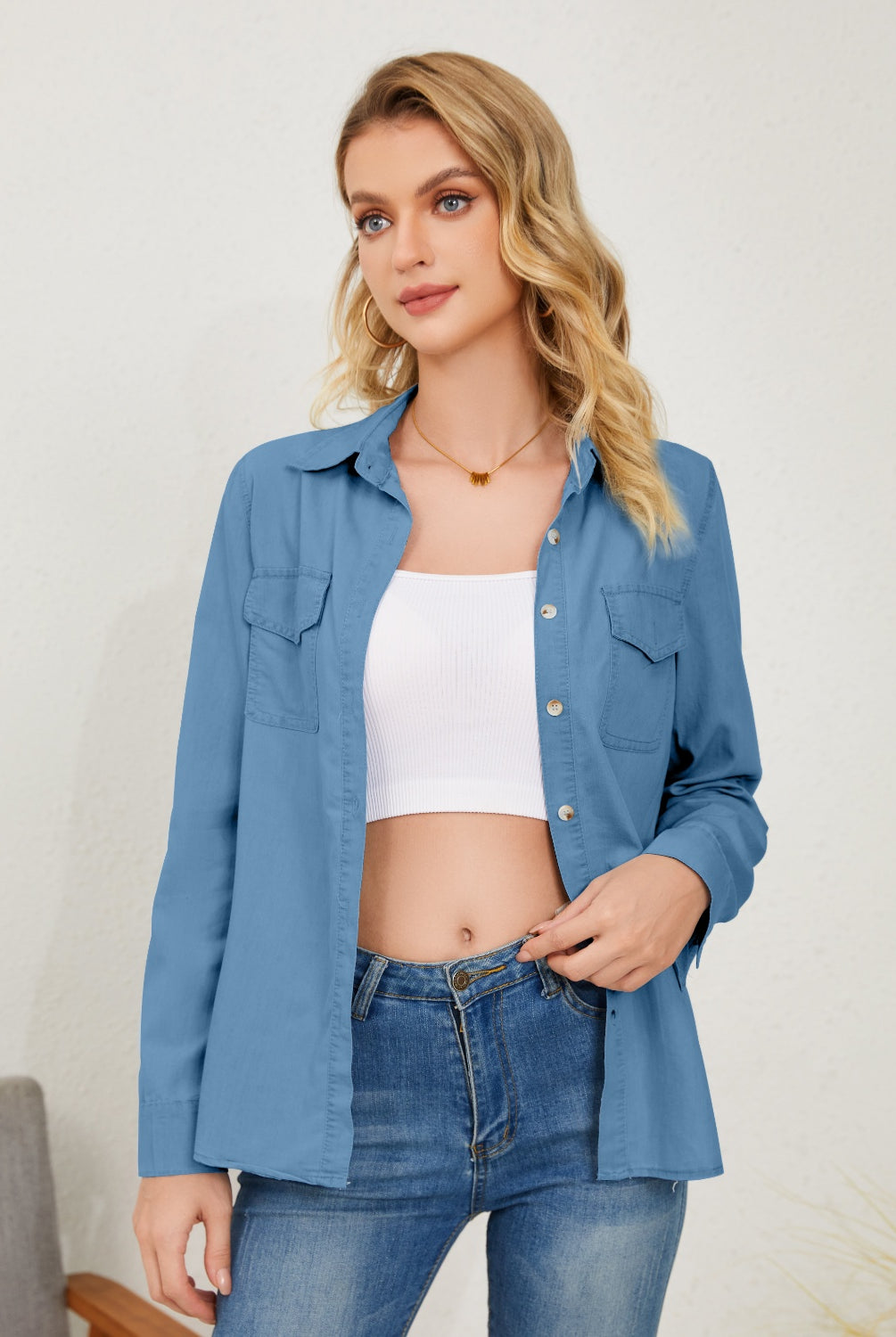 A woman is casually styled in a light denim button-up shirt, left open to reveal a white crop top underneath, and paired with blue jeans for a cohesive, double-denim look.