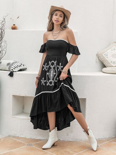 Woman wearing a black off-the-shoulder midi dress with white embroidery, paired with white ankle boots and accessorized with a tan hat and layered necklaces, standing by a white window.
