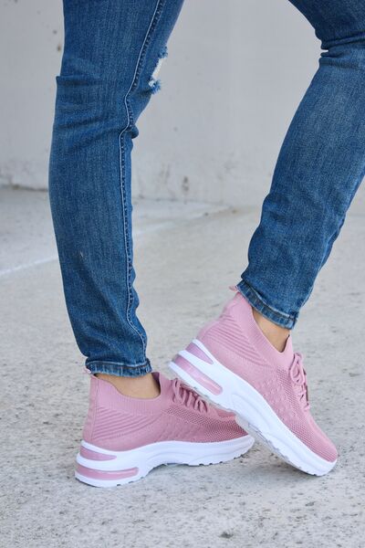 Dusty pink knit athletic shoes with white soles, paired with cuffed blue jeans featuring distressed details, on a concrete background.