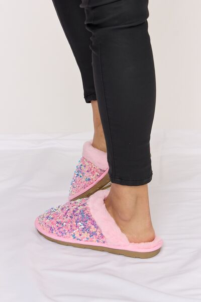 Cozy pink slippers with a multicolored sequin upper and soft faux fur lining, paired with black leggings, on a white satin background.