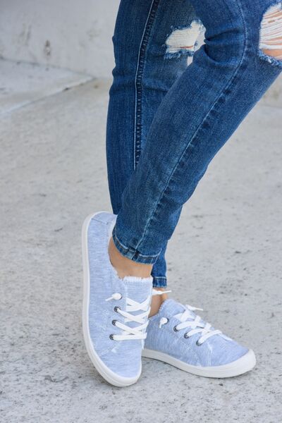 Light blue denim canvas sneakers with white laces and soles, worn with cuffed blue jeans, on a concrete surface.