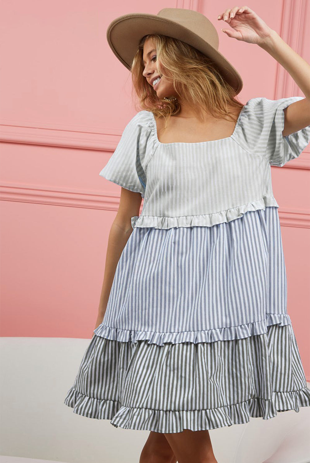 A joyful woman poses in a playful ruffled tiered mini dress with light blue and white vertical stripes, featuring flutter sleeves and a square neckline. She complements the dress with a wide-brimmed tan hat, creating a fresh and fashionable summer look against a pink background.